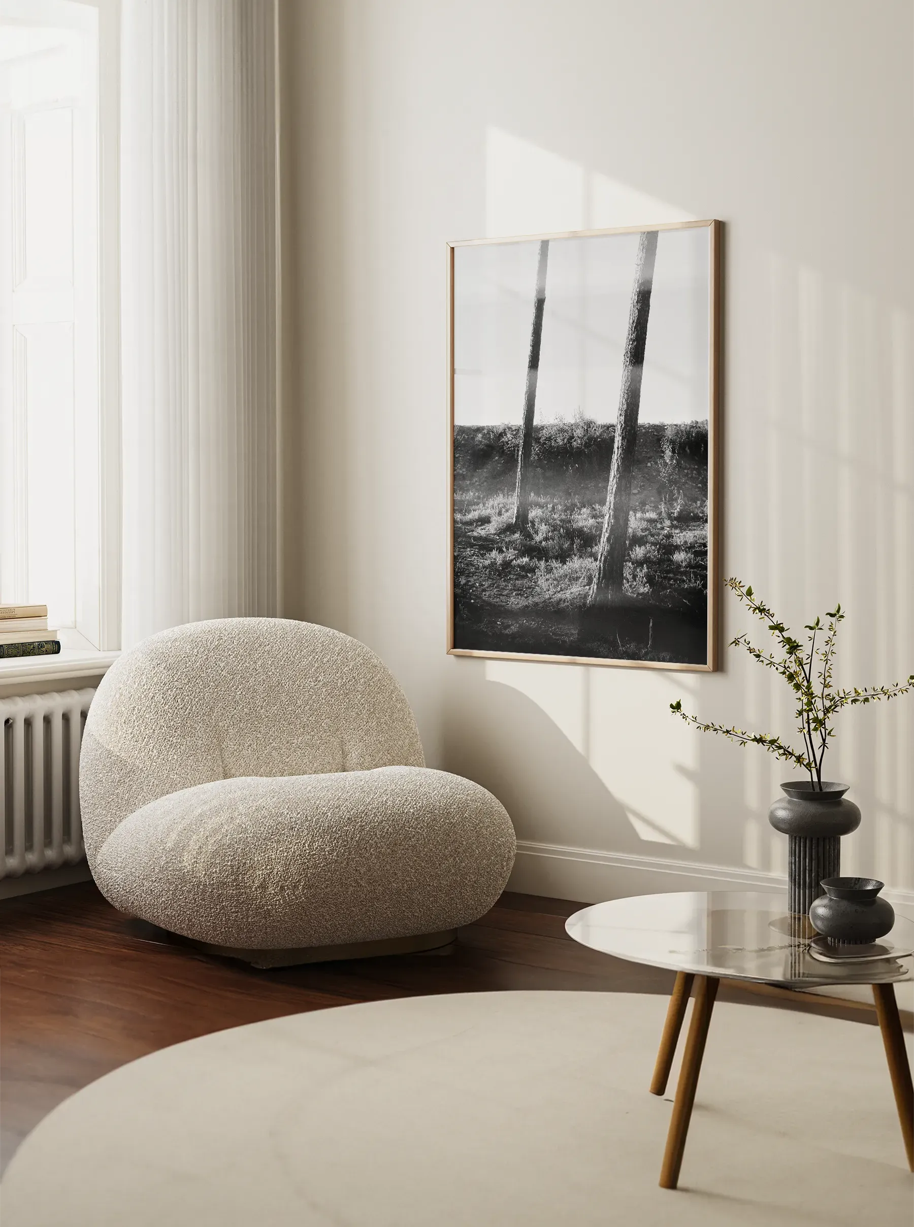 Framed black and white fine art print of a forest scene hanging in a minimalist Scandinavian living room with a soft armchair and round coffee table.