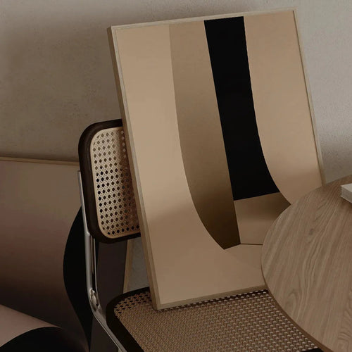 Framed fine art print “Beige Simplicity No.2” by Monday Gallery, styled on a rattan chair beside a wooden table in a minimalist beige interior.