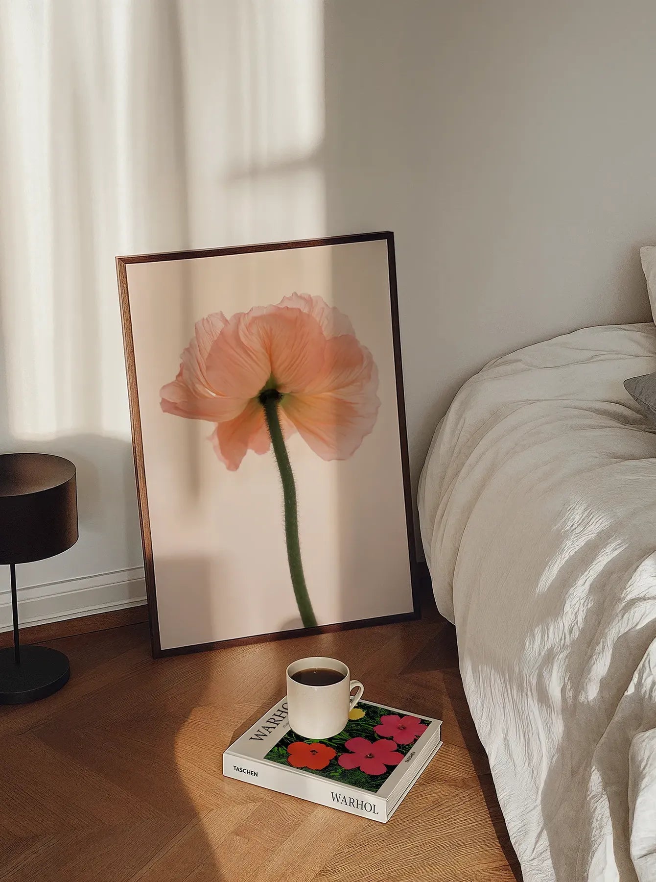 Framed Pink Poppy fine art print leaning against a bedroom wall, styled with soft natural light, a coffee cup, and a Warhol book on a wooden floor. Scandinavian interior design. Printed in Sweden, exclusive at Monday Gallery.